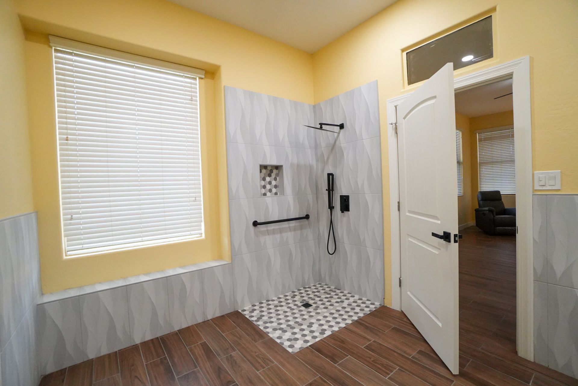 Accessible shower with grab bar and pebble tile floor, next to a room with a recliner.