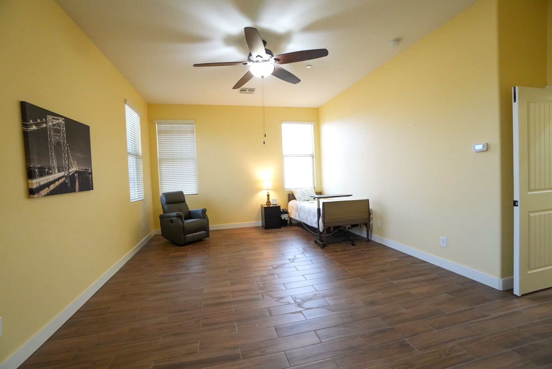 Spacious room with a bed, recliner, and picture. Light yellow walls, wood floor.