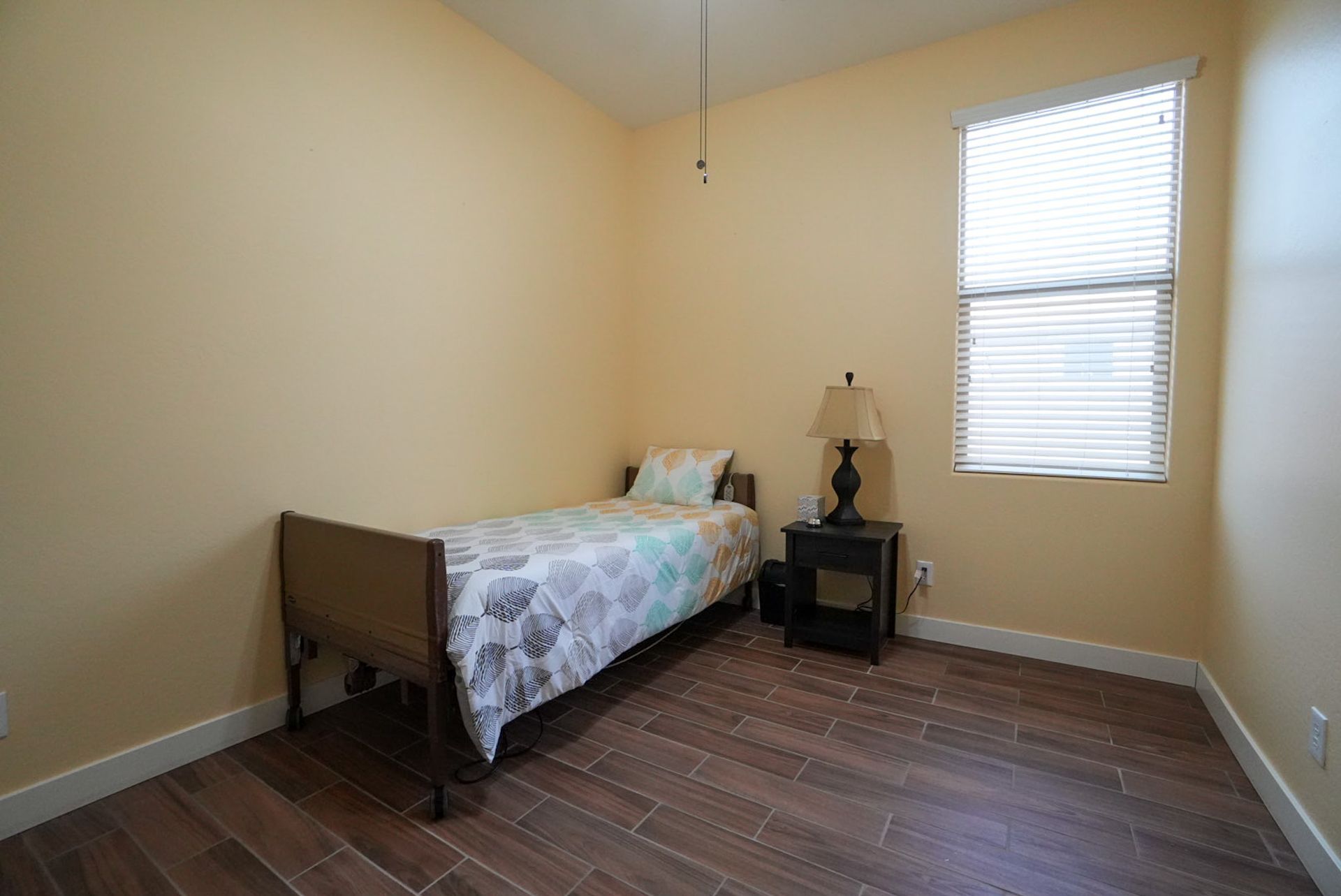 Bedroom with single bed, nightstand, lamp, window with blinds, and wood-look tile floor.