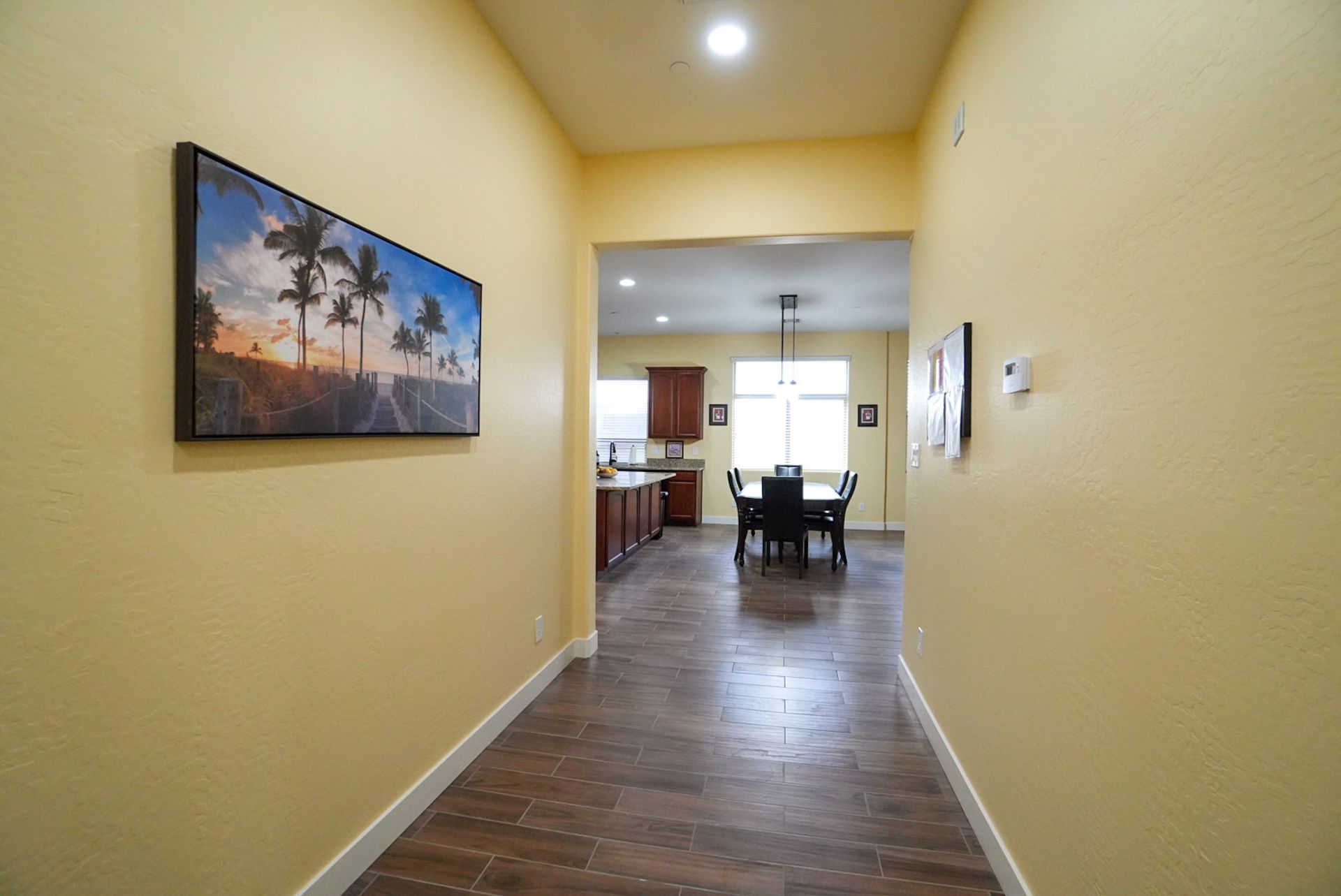 Hallway with artwork, leading to kitchen and dining area. Warm yellow walls and wood floors.