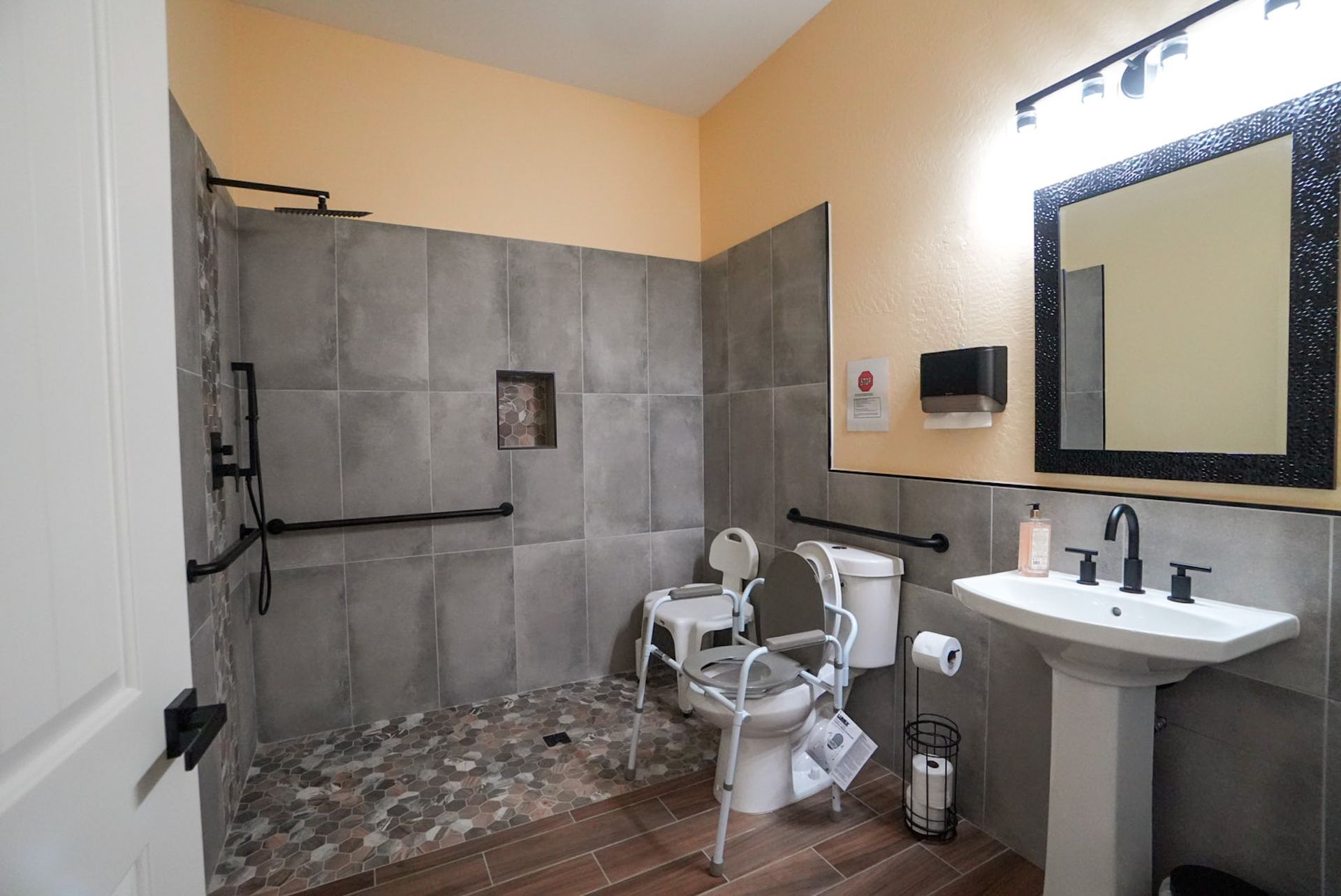 Accessible bathroom with a roll-in shower, toilet, and sink; features grab bars and a shower chair.