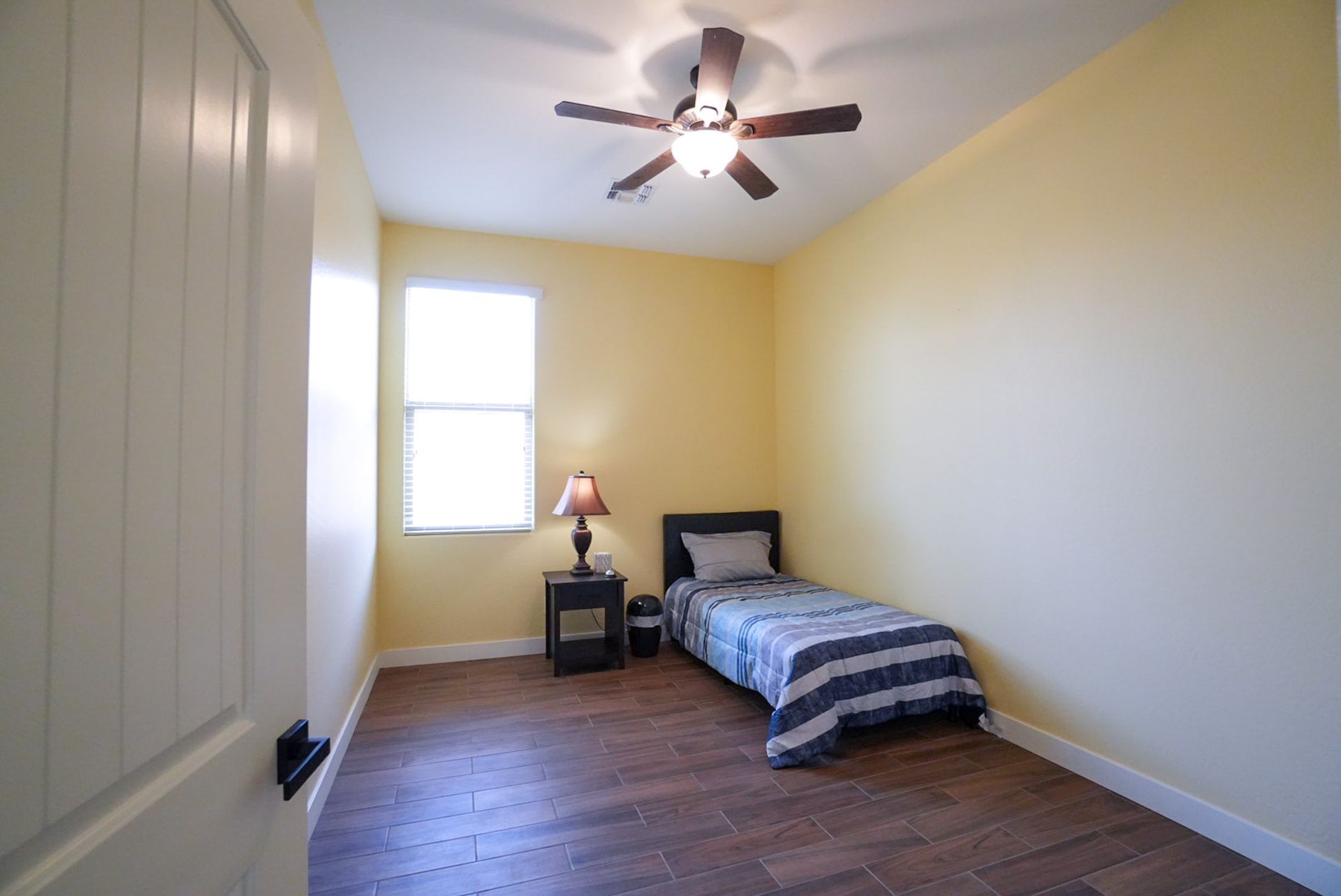 Small bedroom with bed, nightstand, window, and ceiling fan. Yellow walls, wood floor.