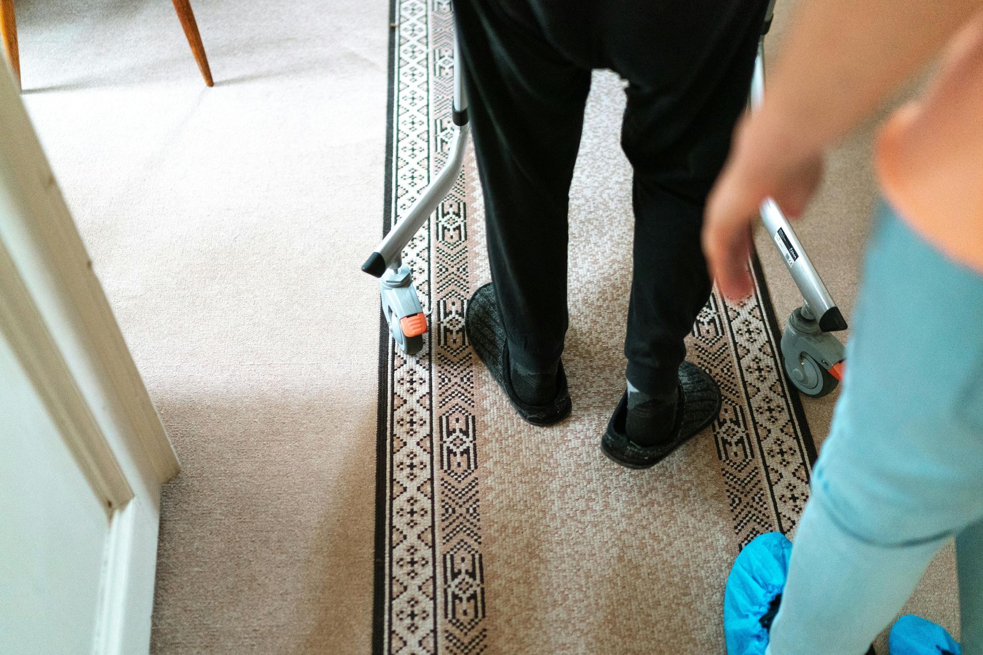 Person using a walker, legs visible, with someone assisting, on a patterned rug in a home.
