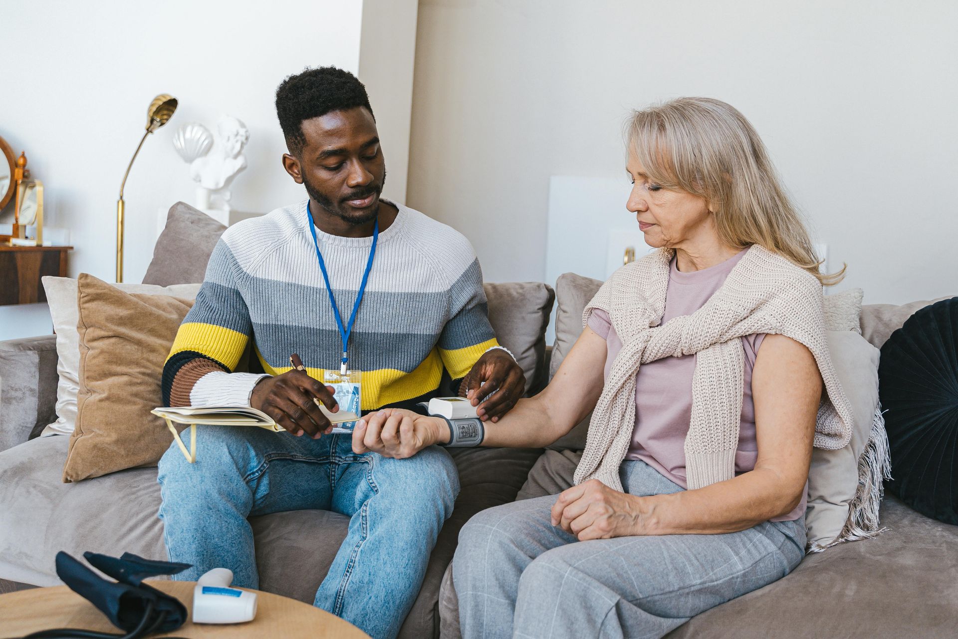 Black man taking an older woman's pulse on a couch. He wears striped sweater and jeans; she has a wrap and gray hair.
