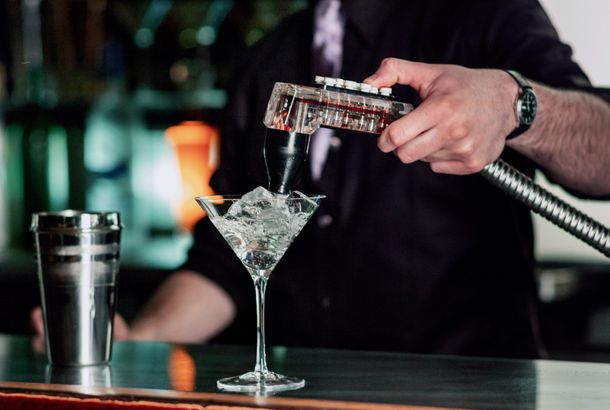 bartender uses fountain soda gun to finish the martini style wine glass filled with ice, near a shaker at a bar counter