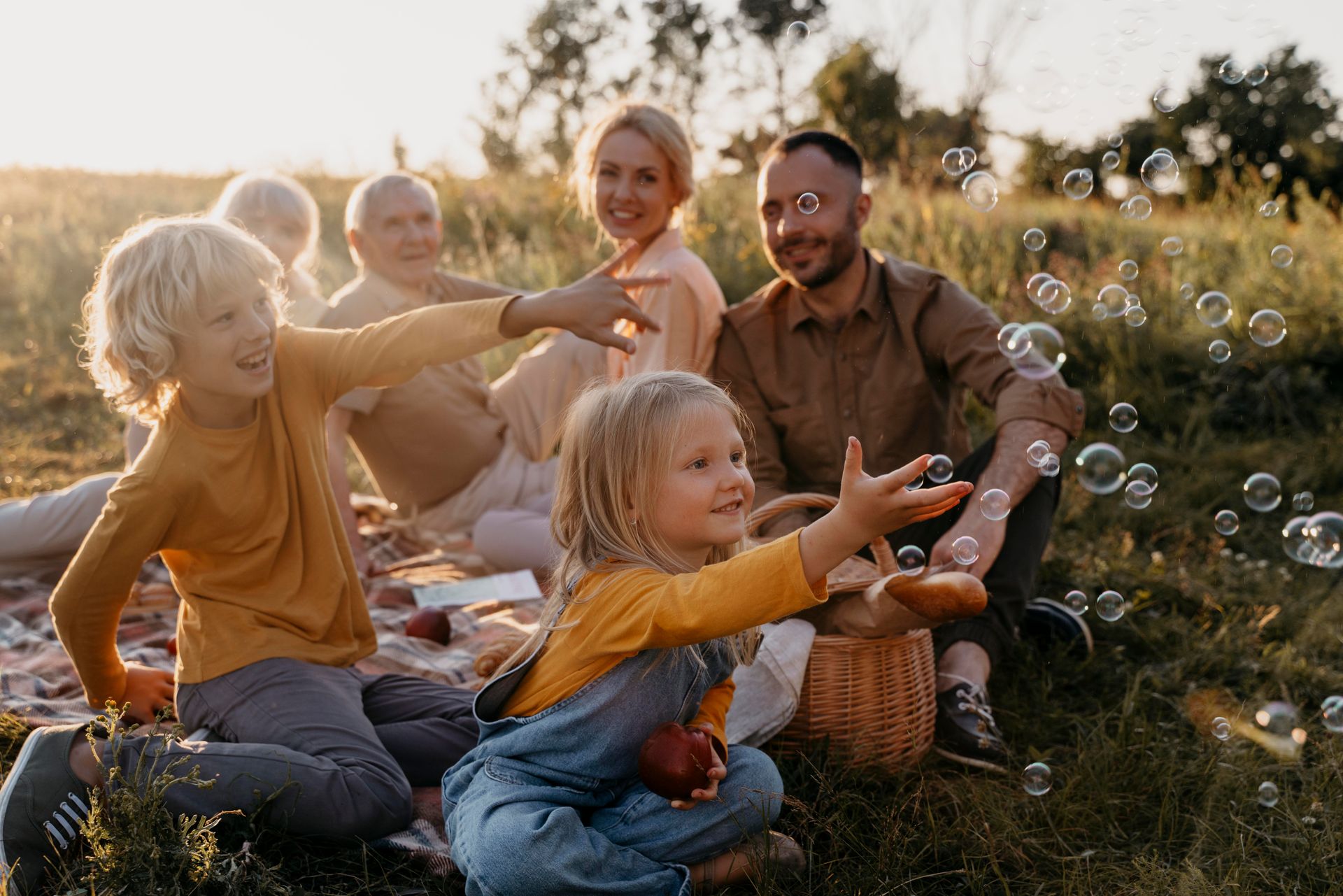 Family picnic in a field, reaching for bubbles. Golden light, blanket, basket.