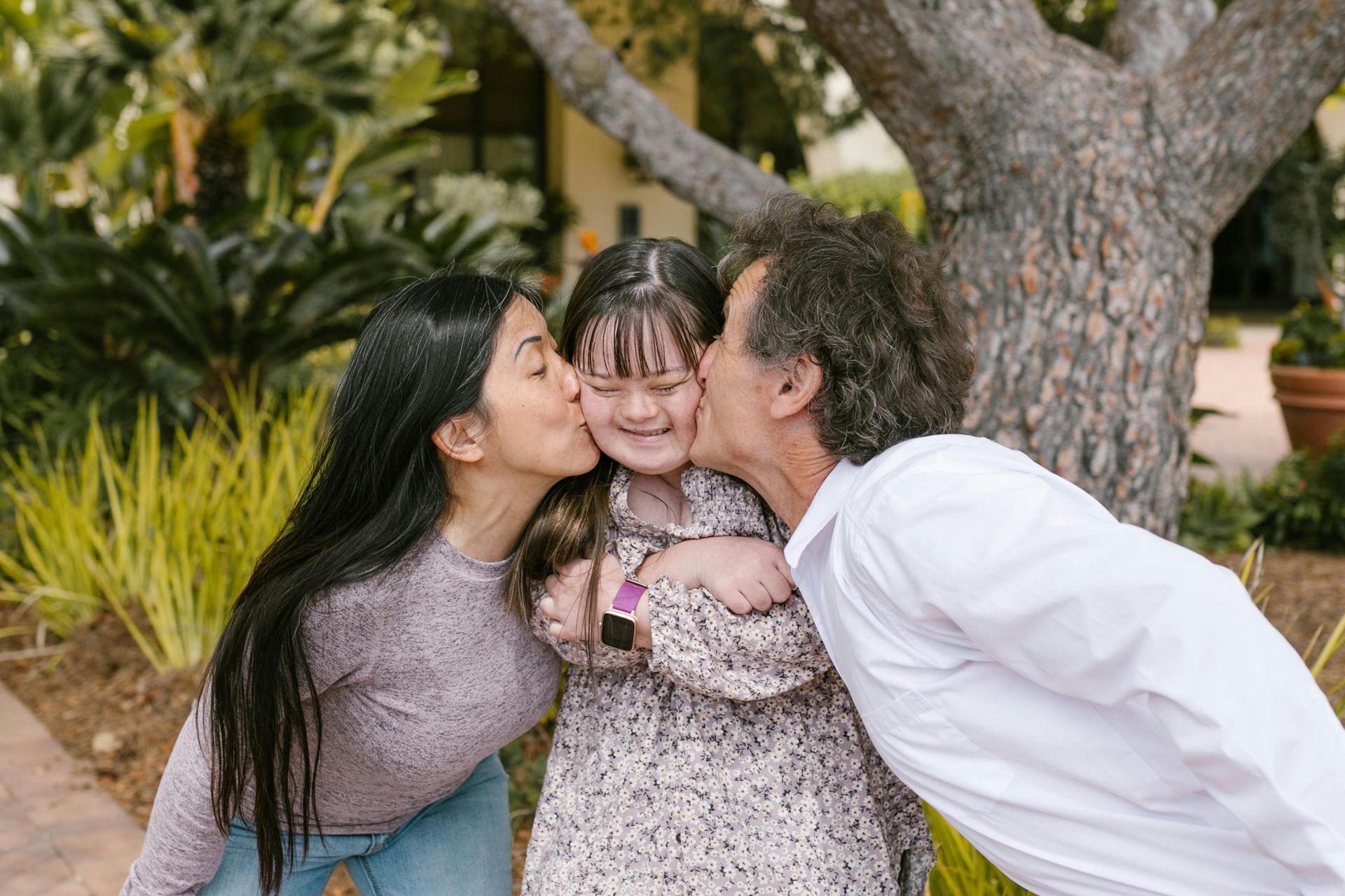 Woman and man kissing a smiling child's cheeks outdoors, embraced near a tree.