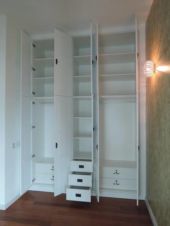 White built-in closet with open doors revealing shelves, drawers, and hanging rods, on a wood floor.