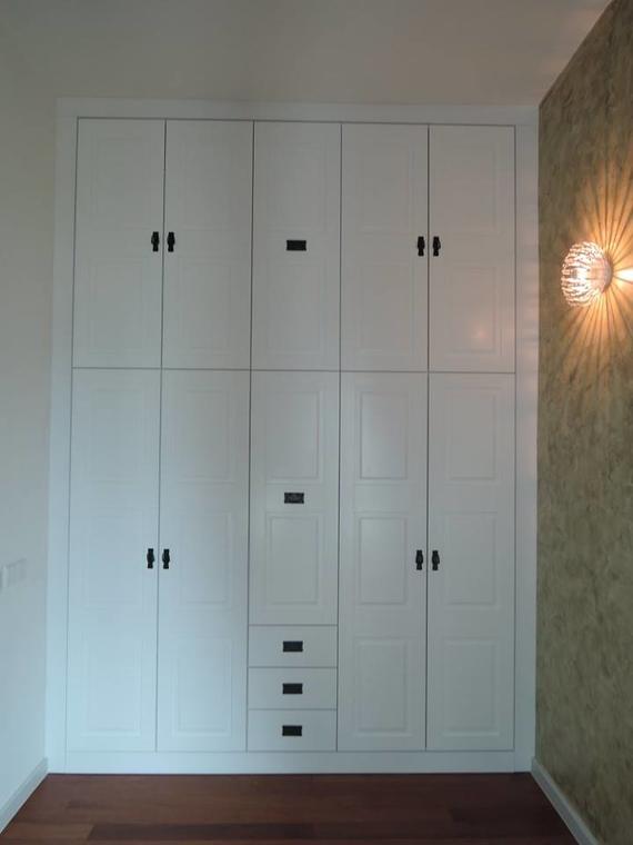 White built-in closet with multiple doors and drawers. Black handles. Dark wood floor. Light fixture on textured wall.