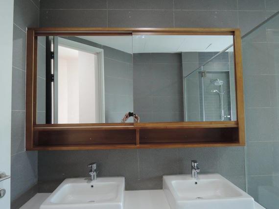 Bathroom with two sinks, large mirror with wooden frame and open shelf. Gray tile walls.