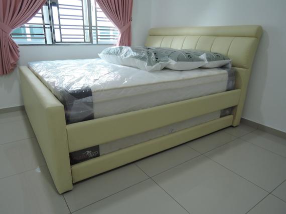 A bed with cream-colored leather frame and headboard, new mattress, and pillows, in a bright room next to a window with pink curtains.