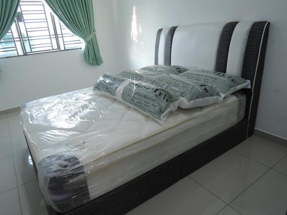 A bed with a white headboard and mattress, pillows, wrapped in plastic in a room with a window.