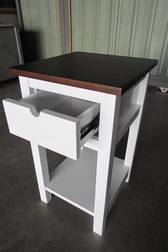 White bedside table with a drawer open, dark brown top, white legs, and a lower shelf.