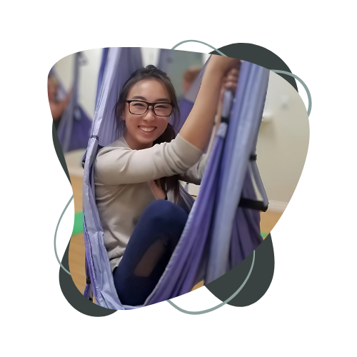 A woman is sitting in a yoga aerial silk hammock and smiling.