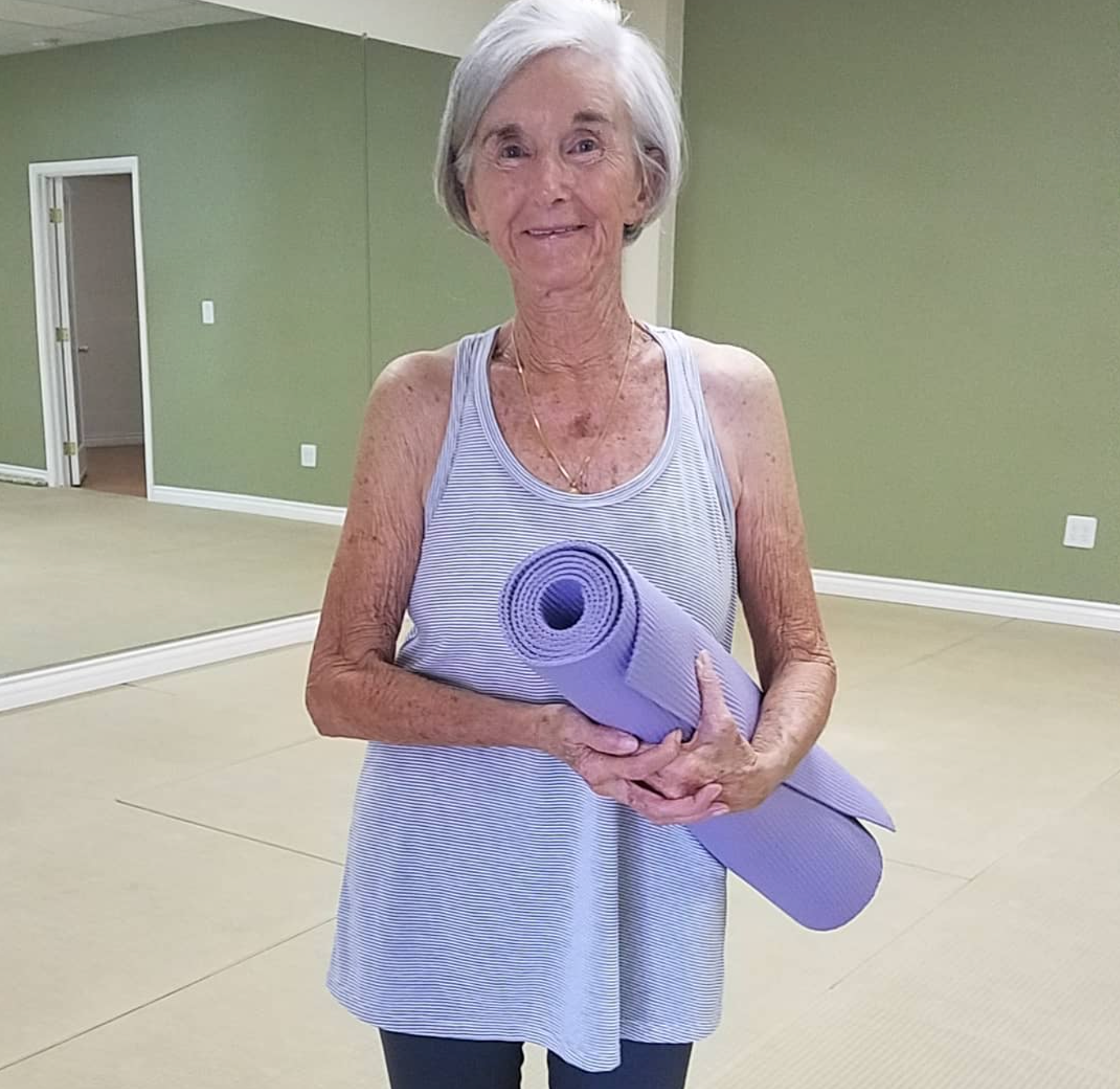 An elderly woman is holding a yoga mat in a gym.