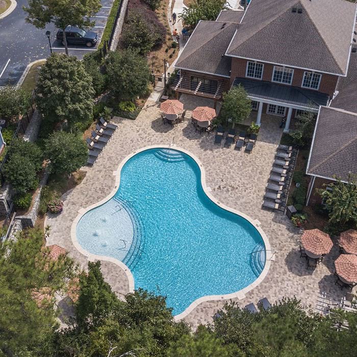 Aerial view of a complex with a large pool, lounge chairs, and an adjacent building with a dark roof.