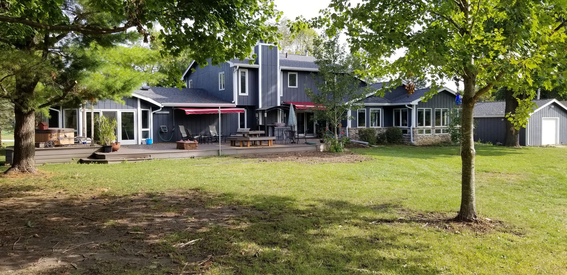 Large gray house with green lawn and trees. Red awnings and patio furniture are visible.