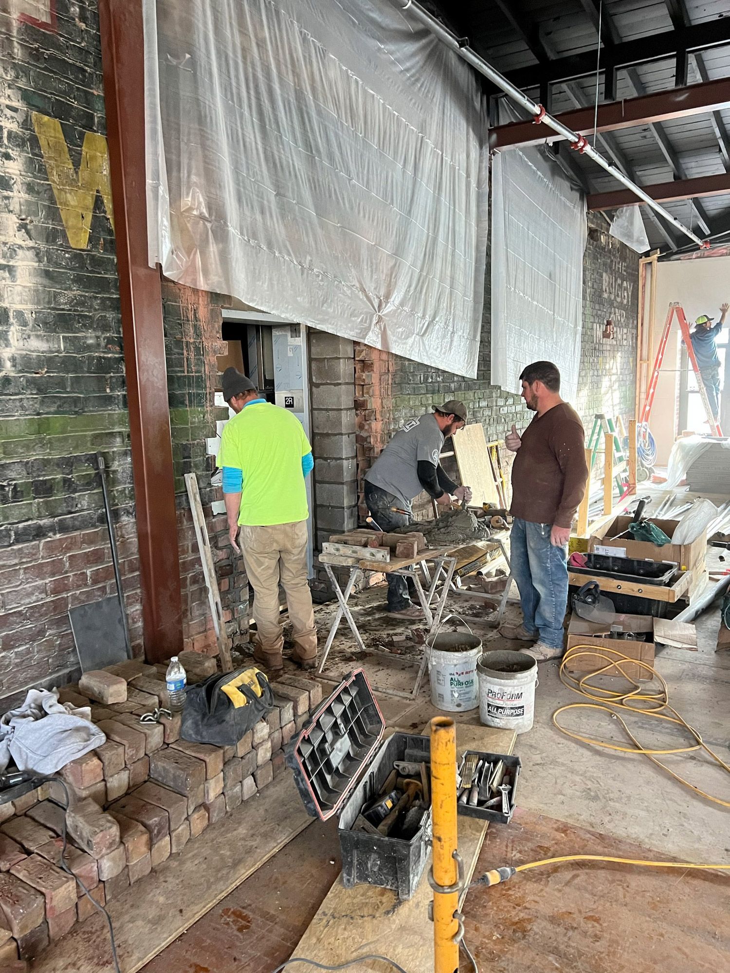Construction workers rebuilding brick wall. Men work indoors near tools, buckets, and support beams.