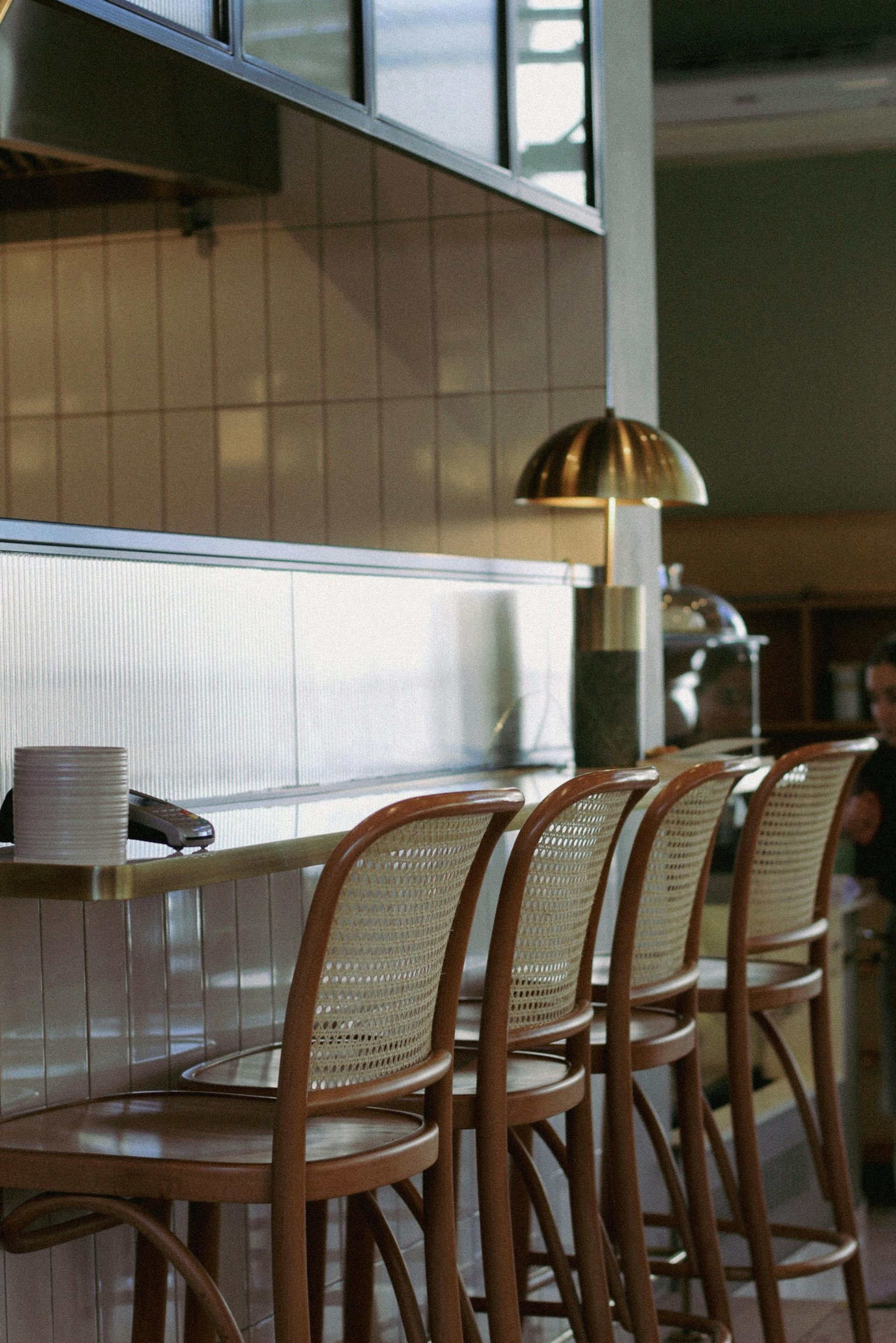Bar stools line a counter in a restaurant, warm wood, and woven backs.