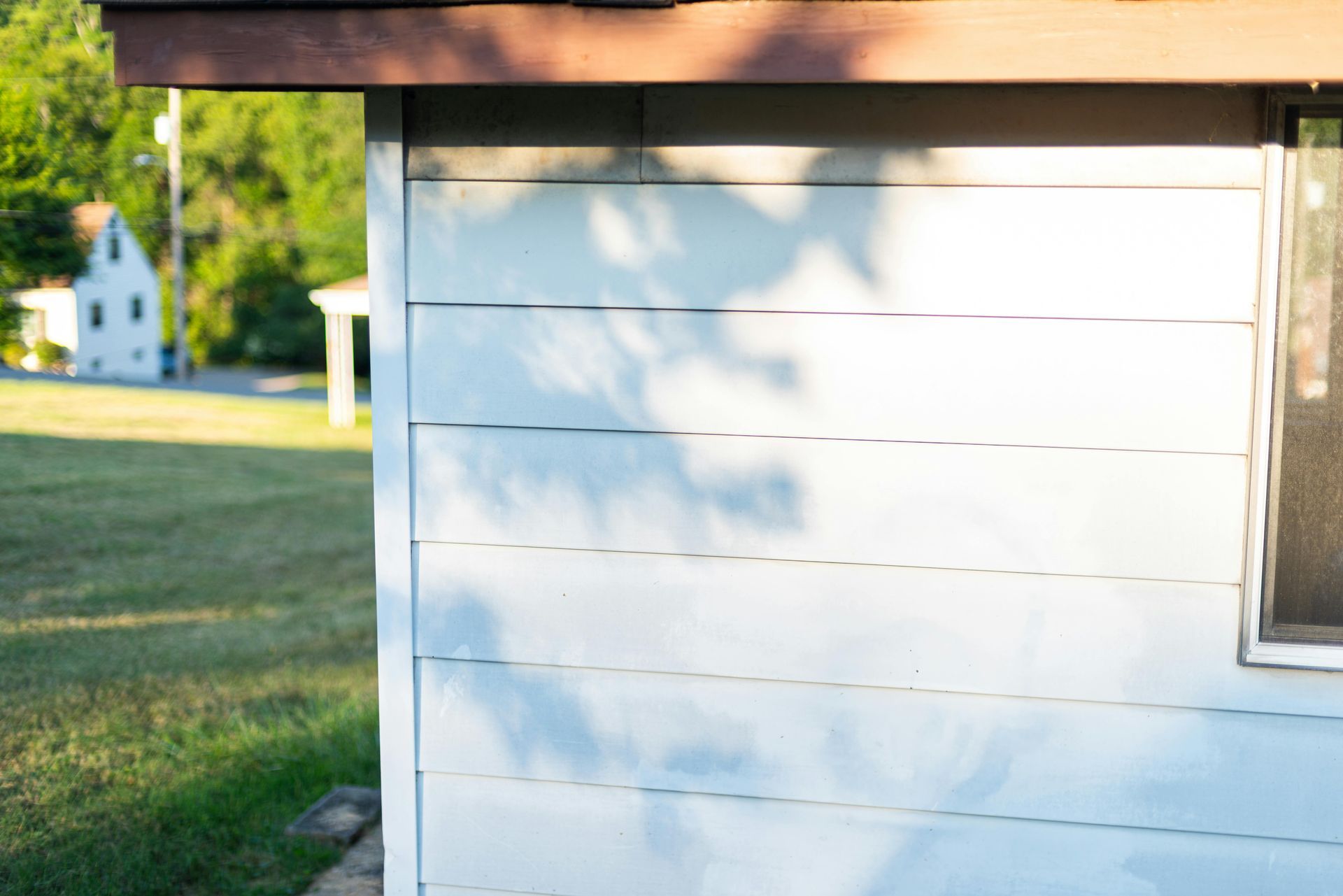 Blue-sided shed with a window, brown trim, and tree shadows, in a yard with a house in the background.