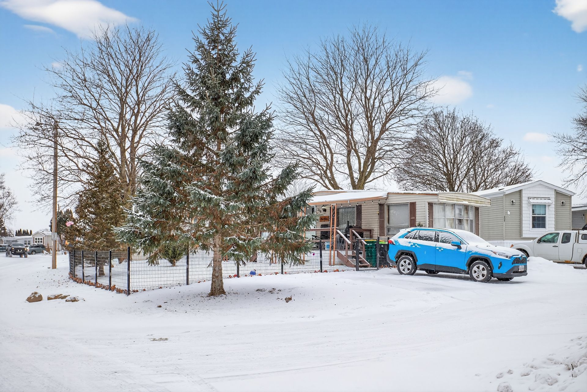 Mobile home in winter setting with snow-covered ground, a blue car, and trees.