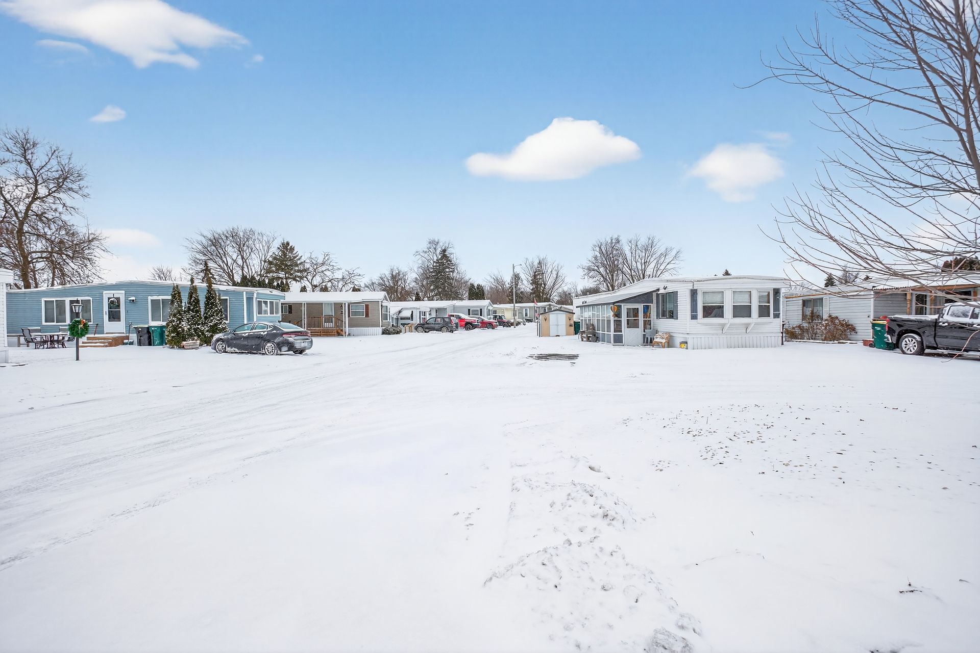 Snowy mobile home park under a blue sky. Cars parked, trees visible.