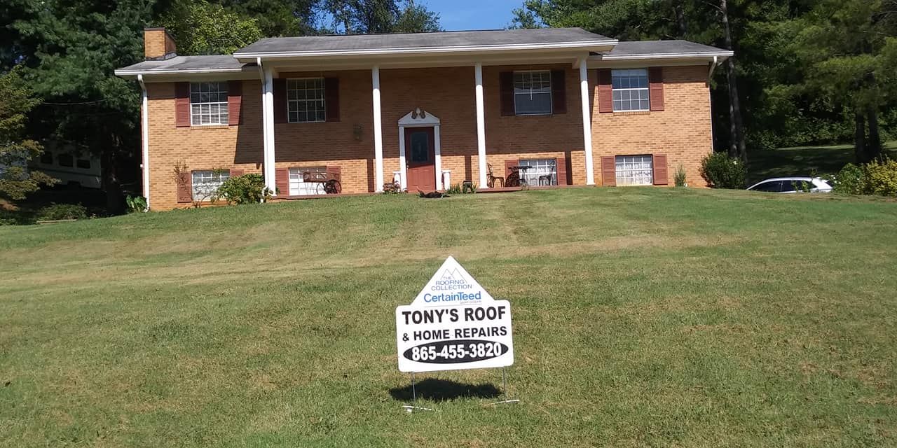 A brick house with a sign in front for Tony's Roof. The house is on a grassy hill.