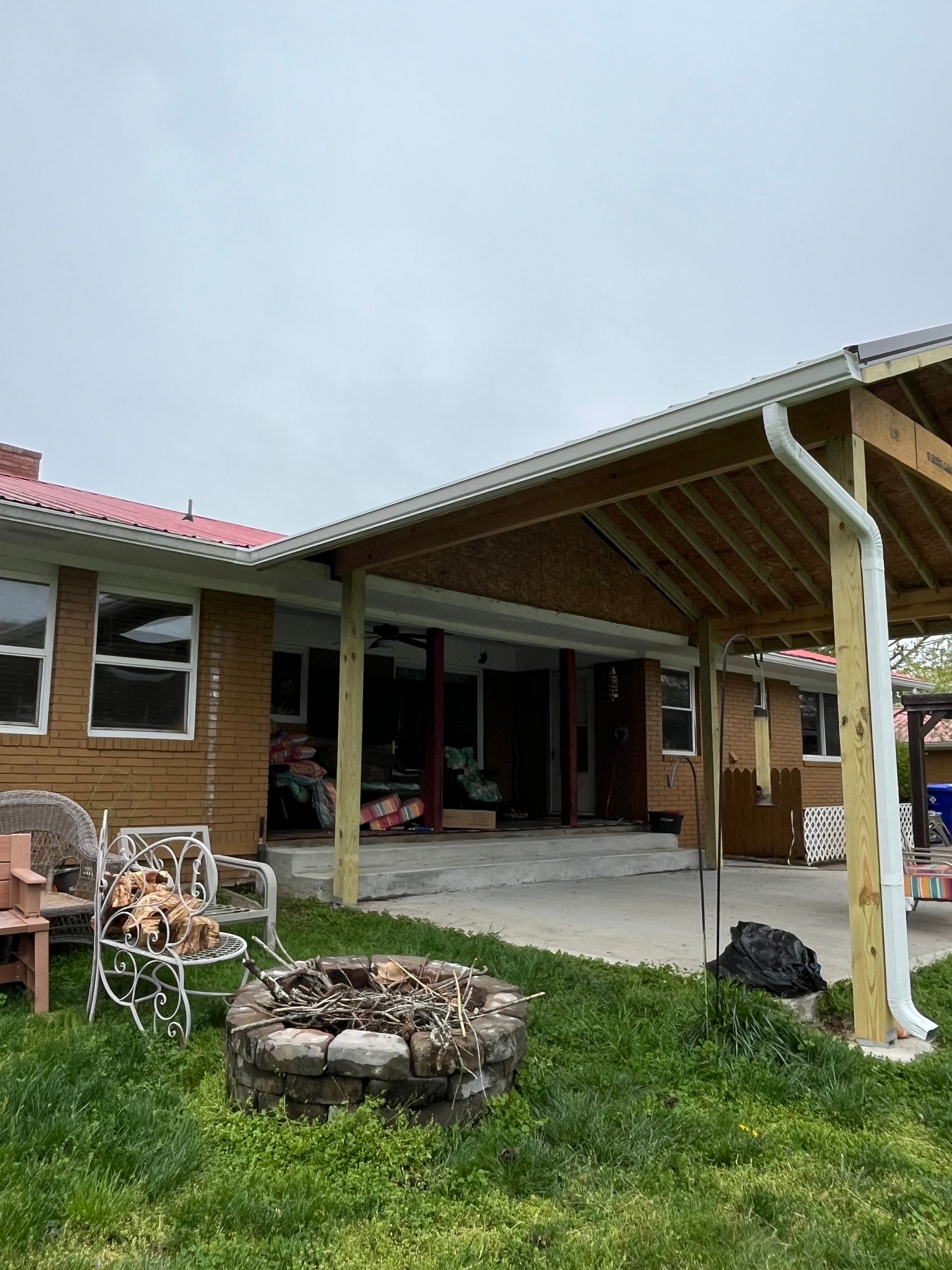 Backyard with a patio, fire pit, and covered porch. The house is brown brick with a red roof. Cloudy sky.
