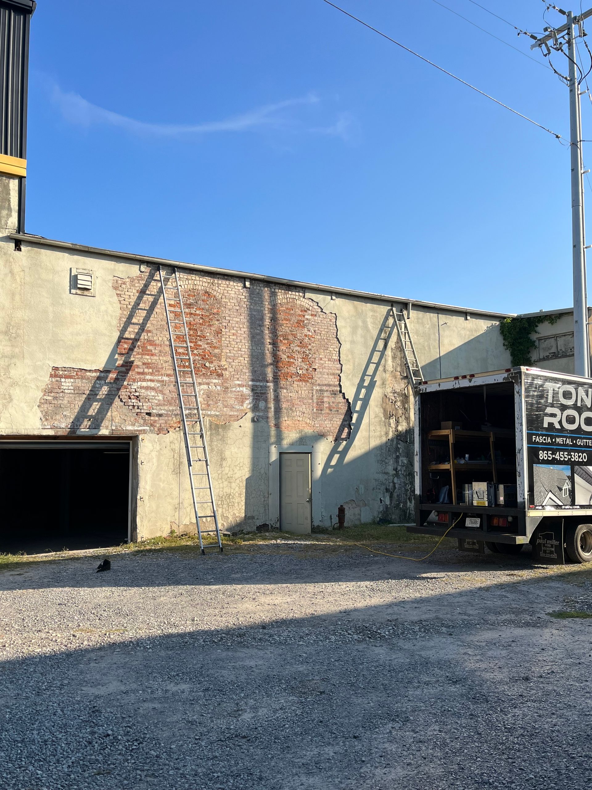 Exterior wall with exposed brick patches, ladders, and a truck parked on gravel.