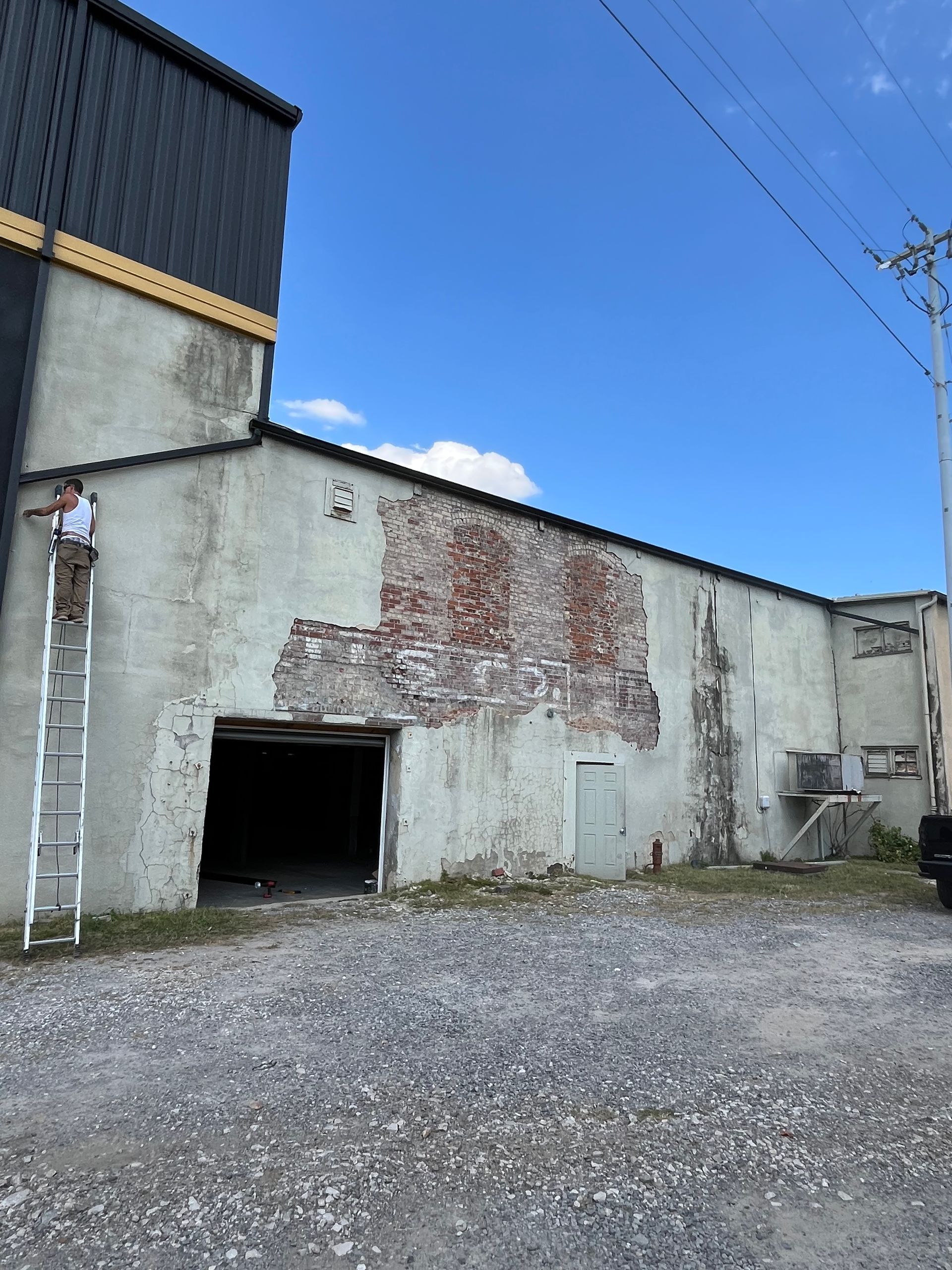Side of a weathered brick building with a large opening, ladder, and person near a sky.