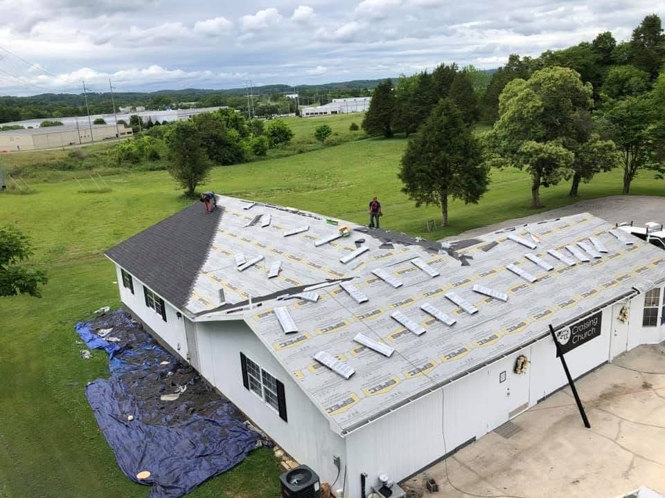 House with roof partially covered with roofing material, a worker standing, blue tarp on ground.