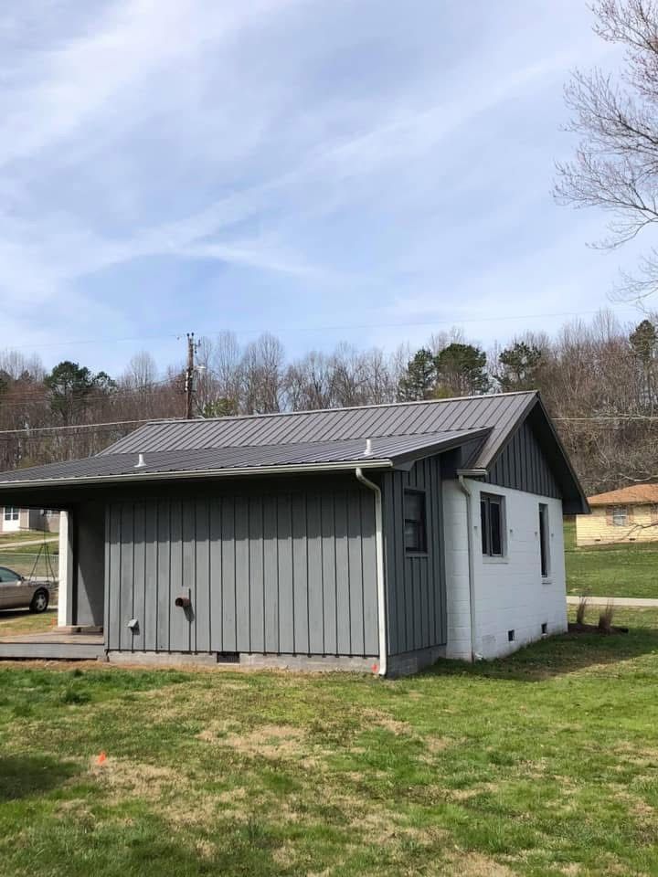 Gray house with metal roof, partially covered porch, and white side; set on green lawn.