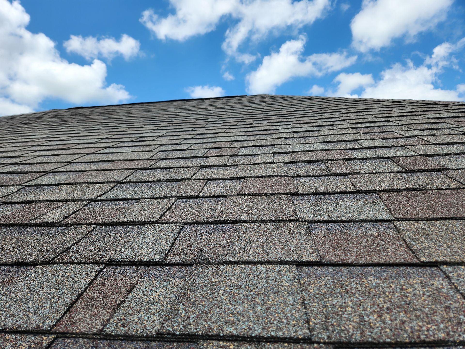 Close-up of a brown asphalt shingle roof against a partly cloudy blue sky.