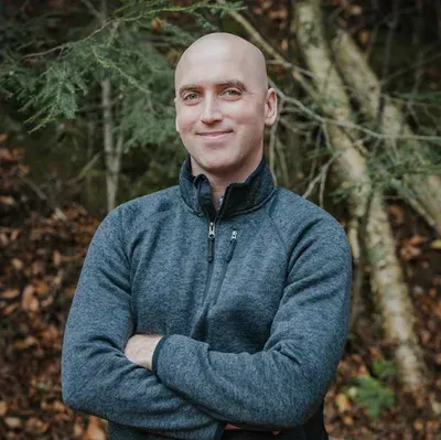 A man is standing in the woods with his arms crossed and smiling