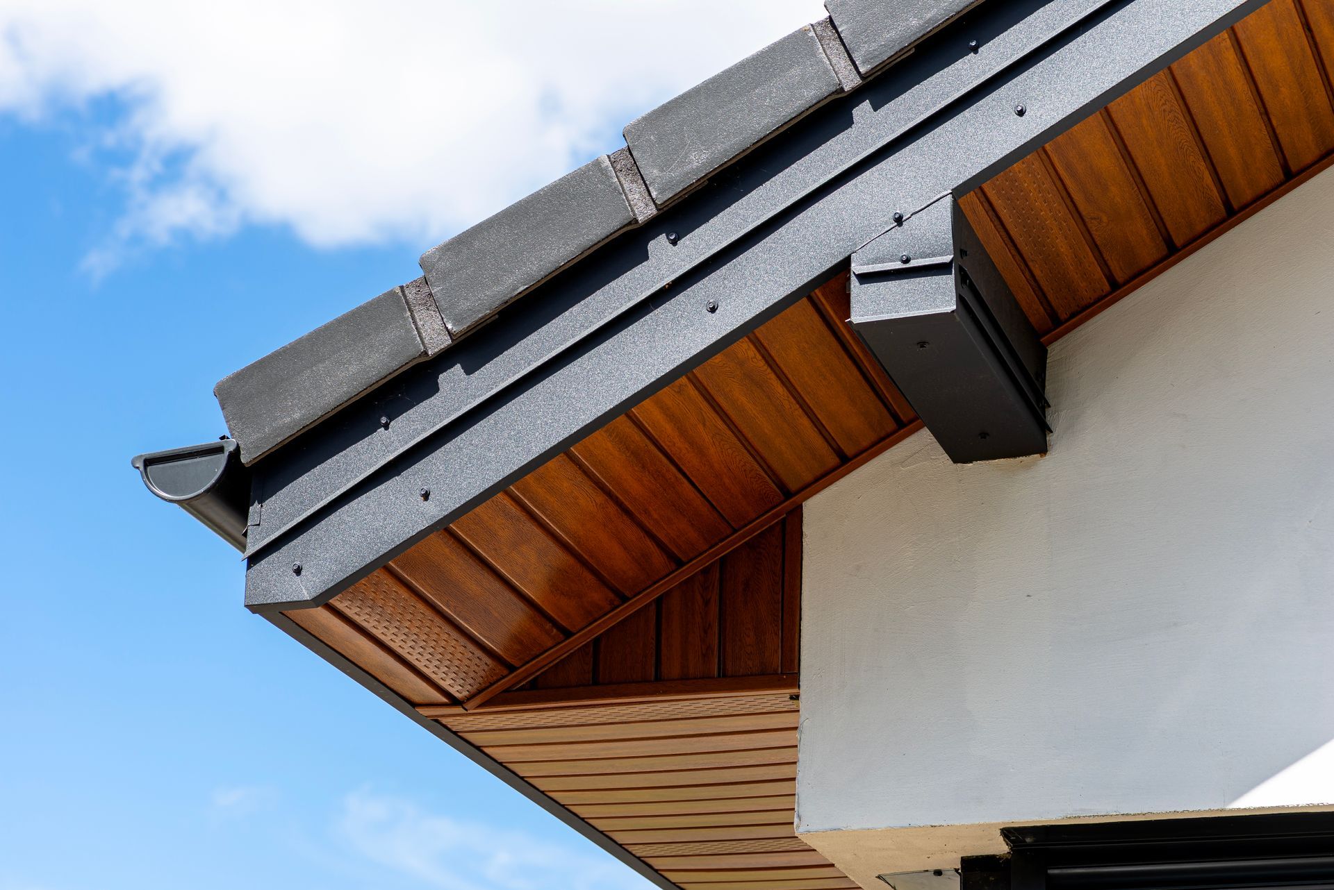 Close-up of a house roof eave with gray shingles, black gutters, and wood soffit against a blue sky