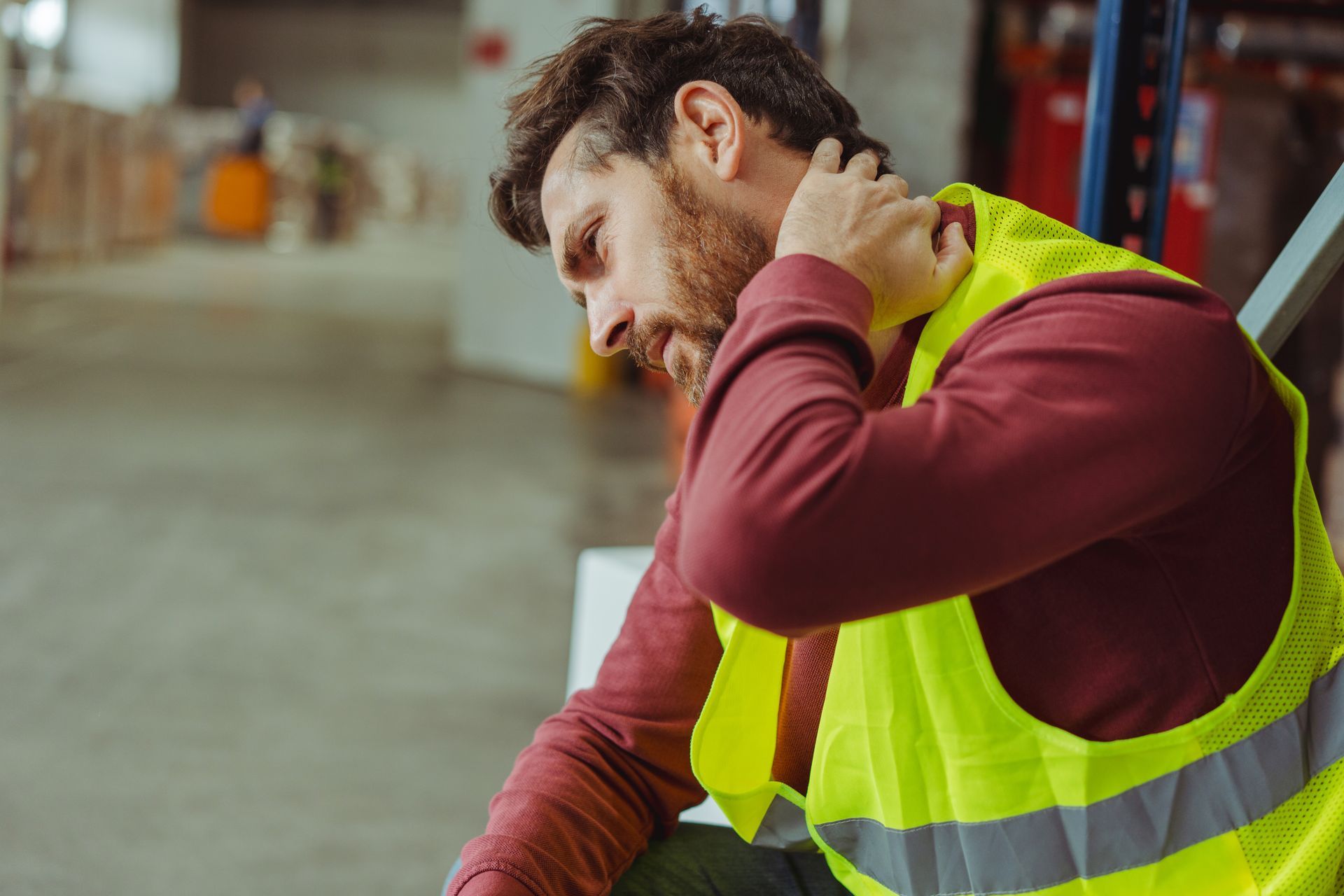 Warehouse worker in safety vest holding neck, looking down, in pain.