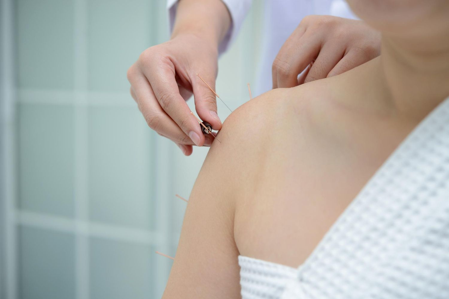 Person receiving acupuncture on shoulder. Needles inserted by hands. White robe and setting.