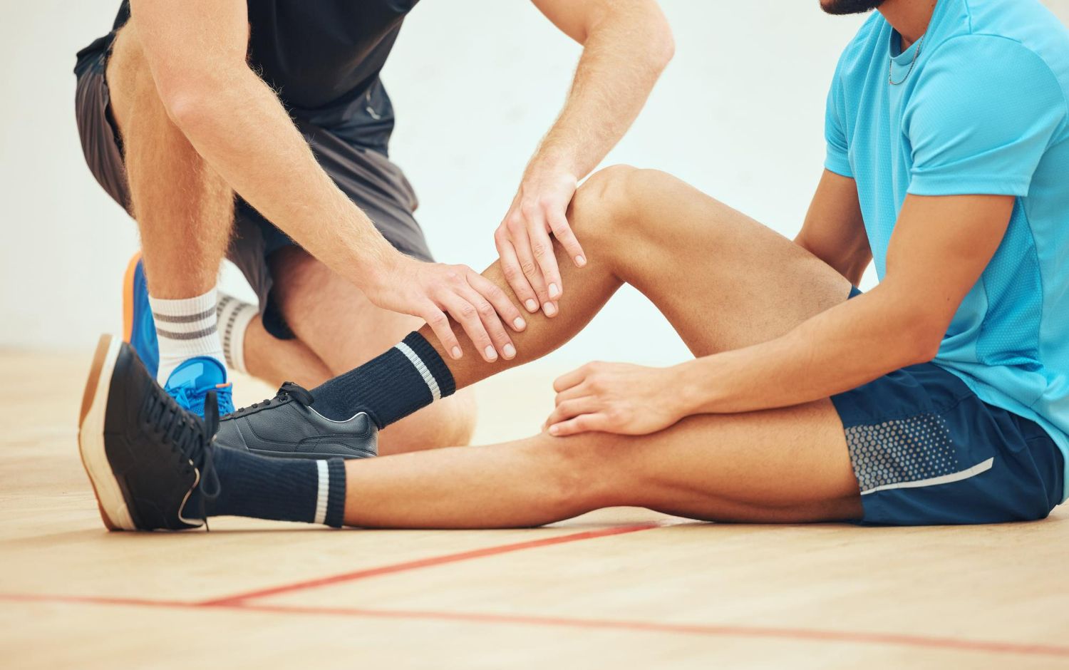 Man seated on floor, leg injury examined by another.  Indoor setting, both wearing athletic clothing.