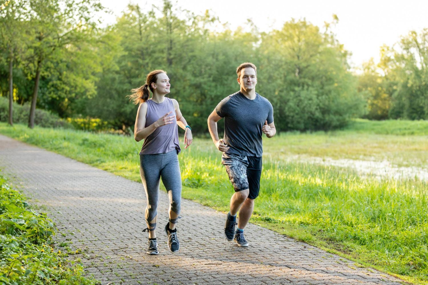 Two people running on a paved path in a park; sunny, green background.