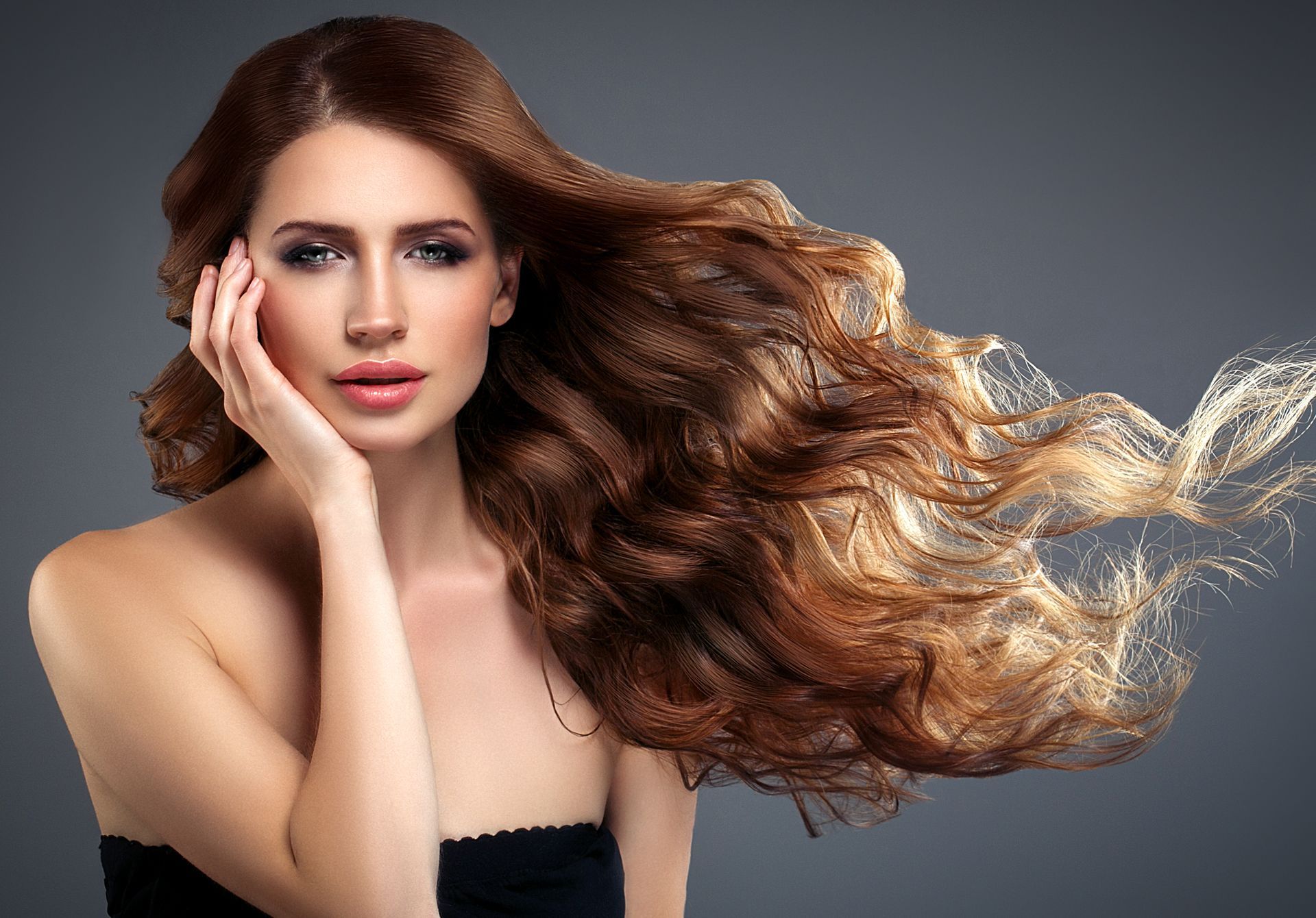 Woman with flowing auburn hair, hand on cheek, against a gray background.