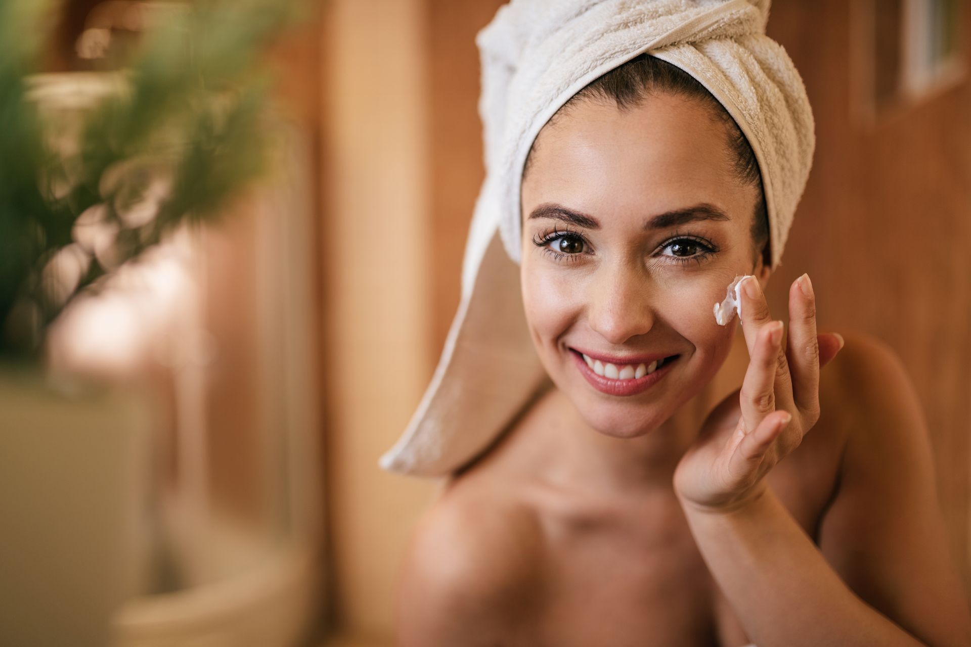 A woman with a towel wrapped around her head is applying lotion to her face.