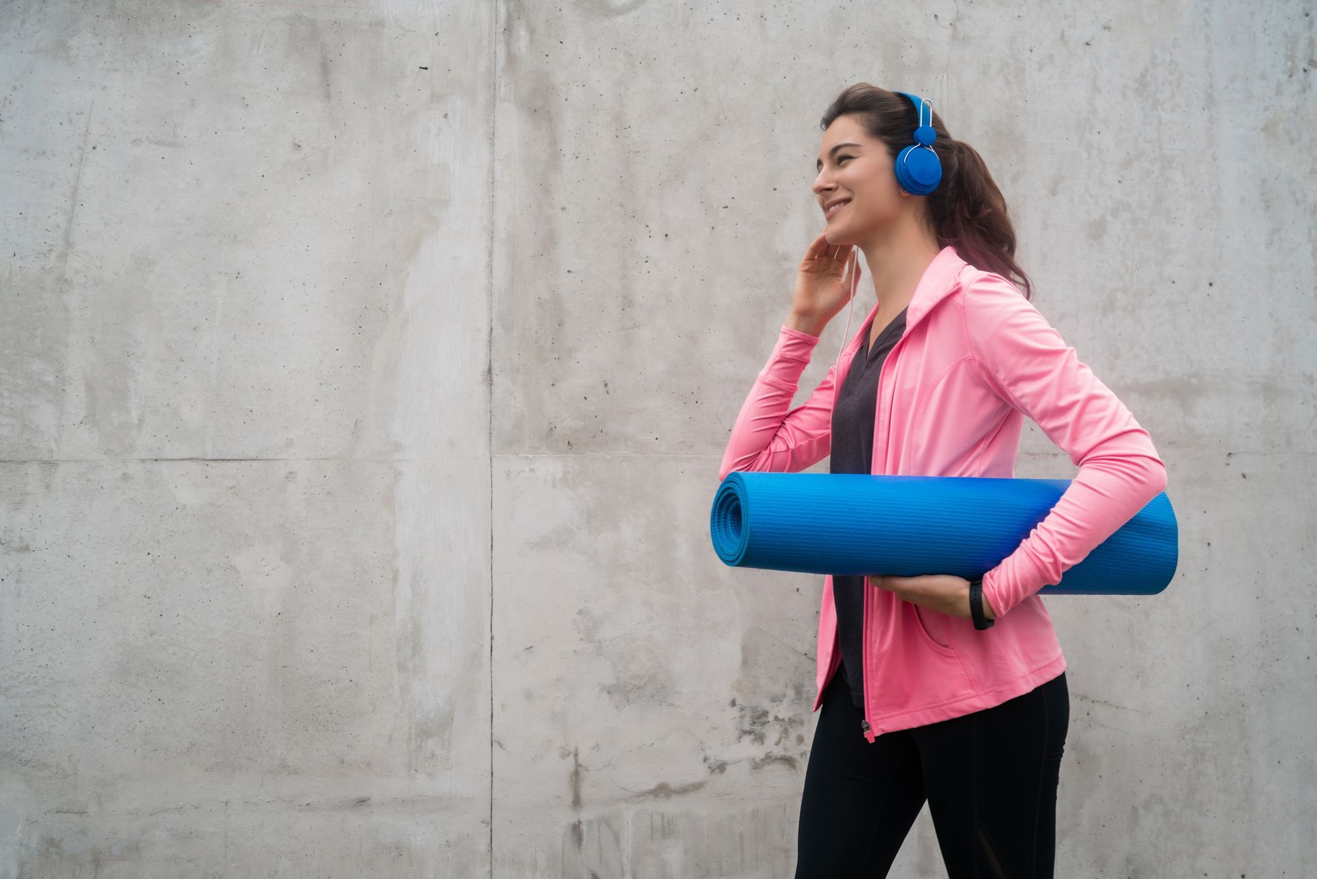 A woman wearing headphones is carrying a yoga mat and listening to music.