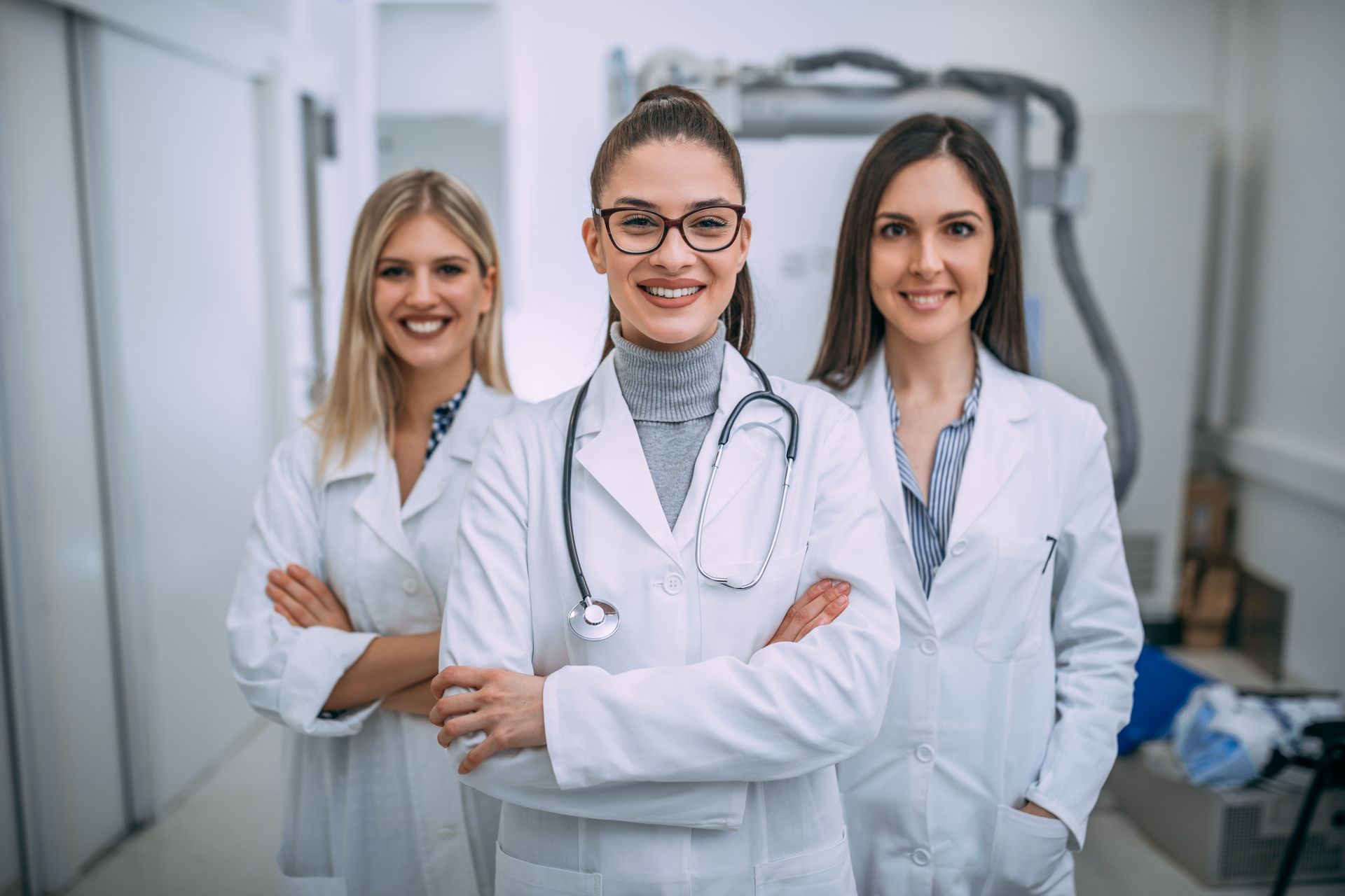 Three female doctors in white coats standing arm in arm, smiling in a medical hallway.