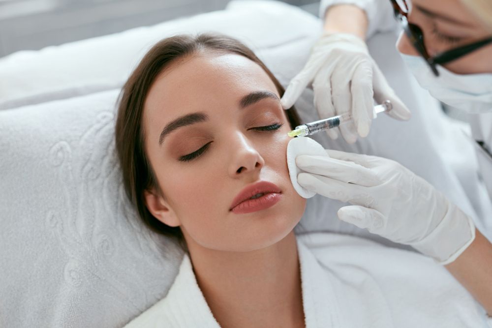 Woman receiving a cosmetic injection near her eye while lying down. A professional in gloves and a mask administers the injection.
