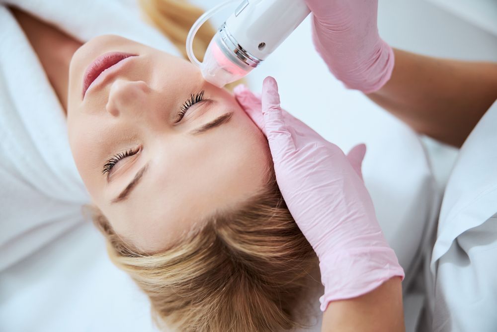 Woman receiving facial treatment with a laser device, held by gloved hands.