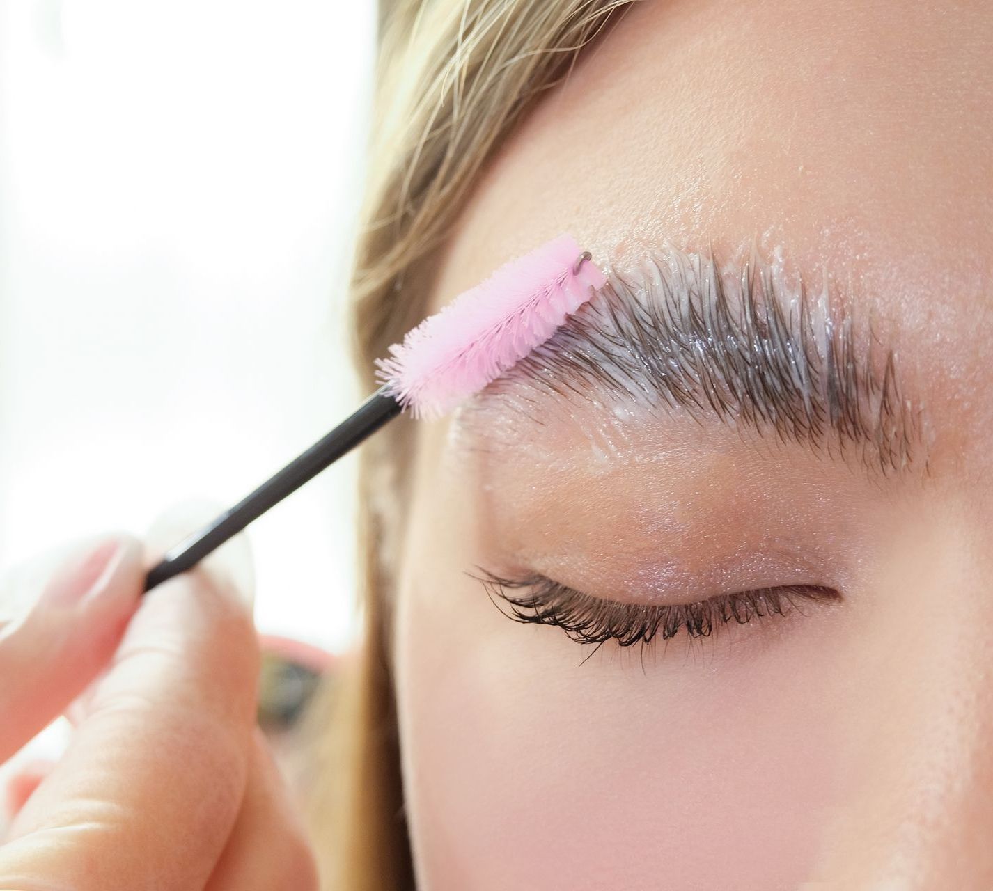 Person's eyebrow being styled with a pink brow brush. White product is applied and smoothed through the eyebrow.