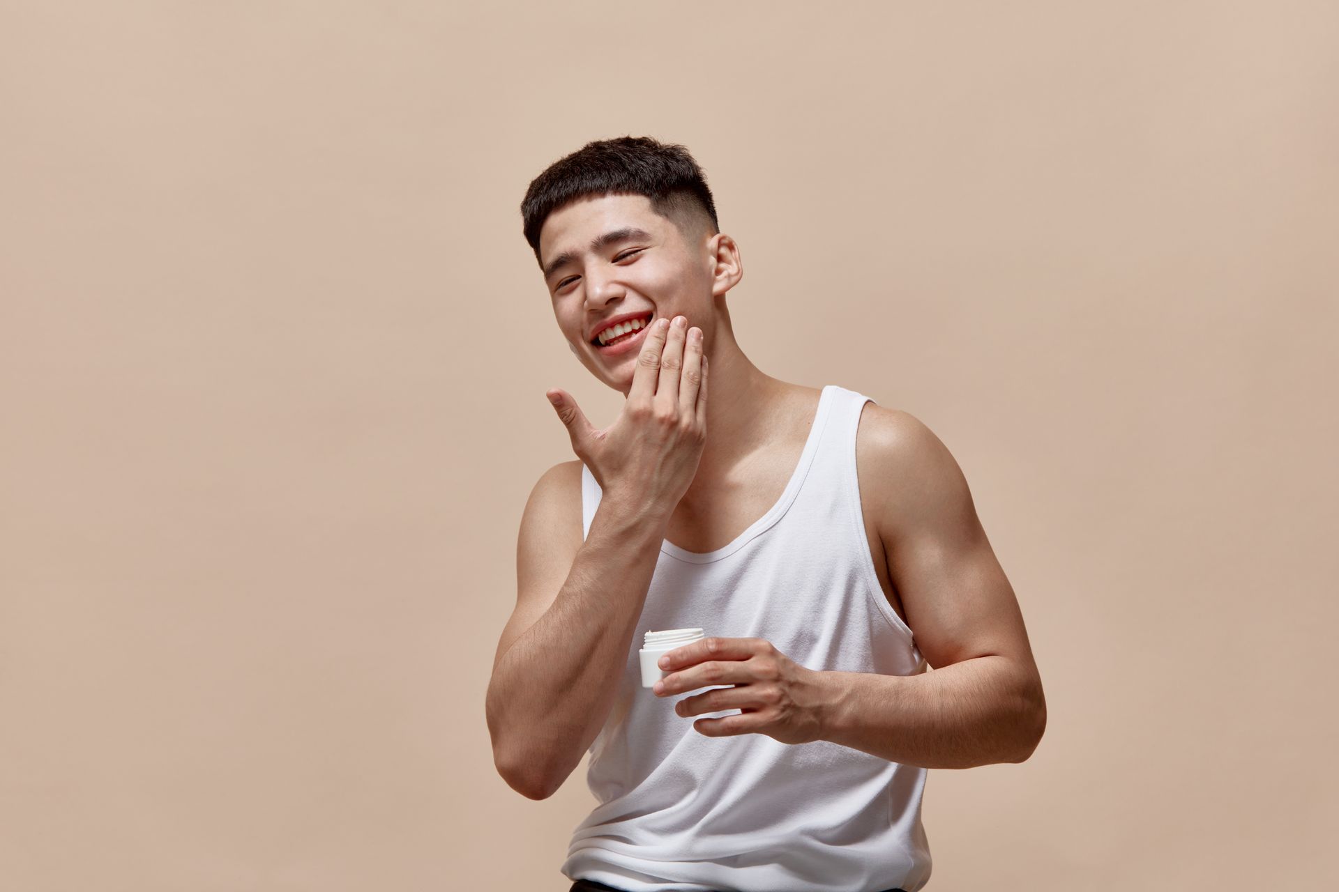 A young man in a white tank top smiles while applying something to his face, set against a neutral background.