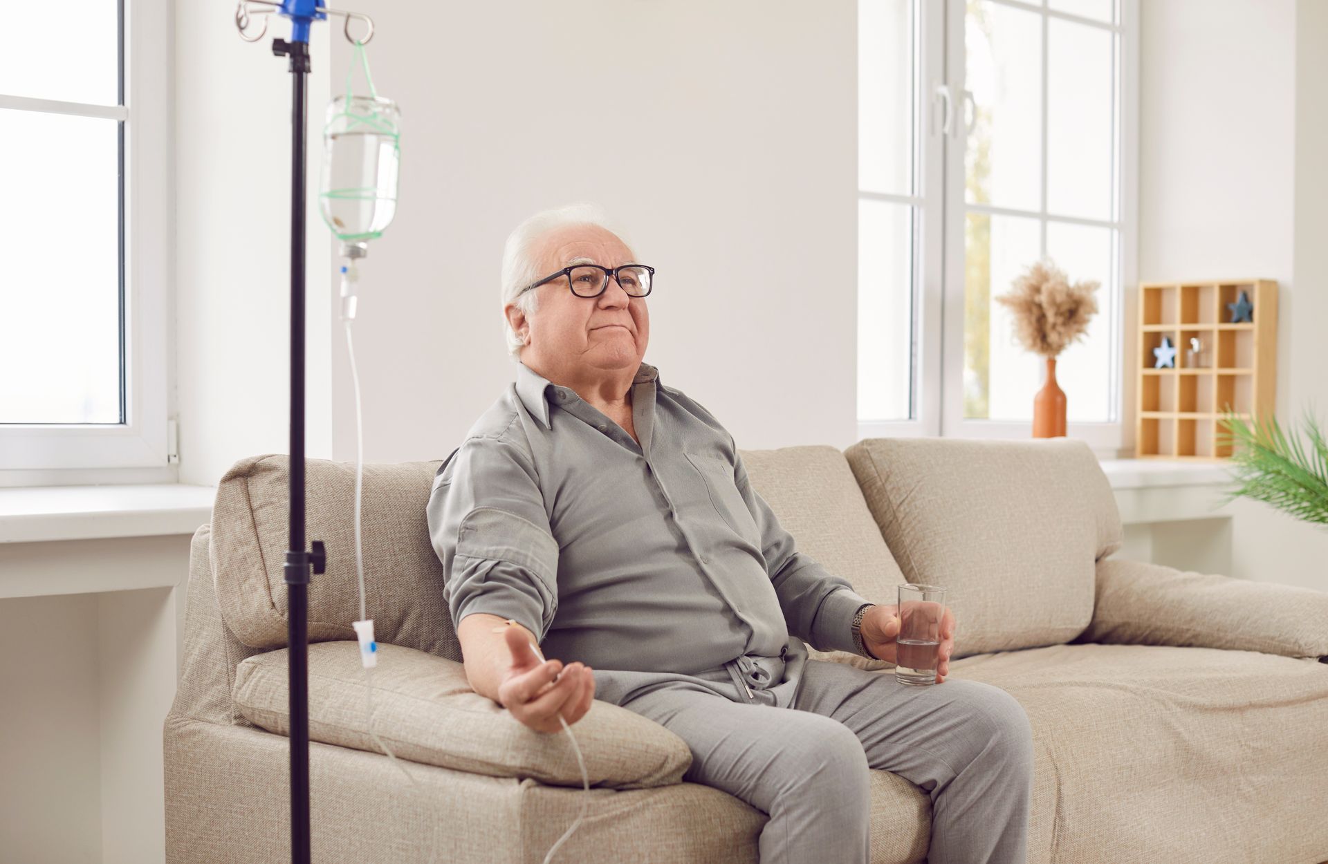 Elderly man receiving IV fluids while seated on a couch near a window. He wears glasses and looks upward.