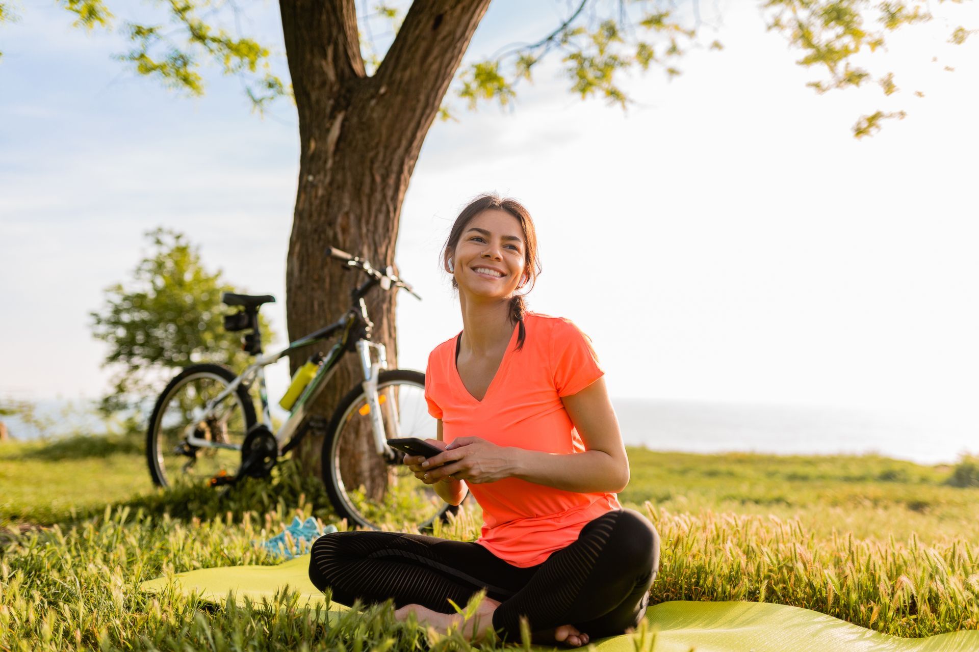 A woman is sitting on a yoga mat under a tree next to a bicycle.