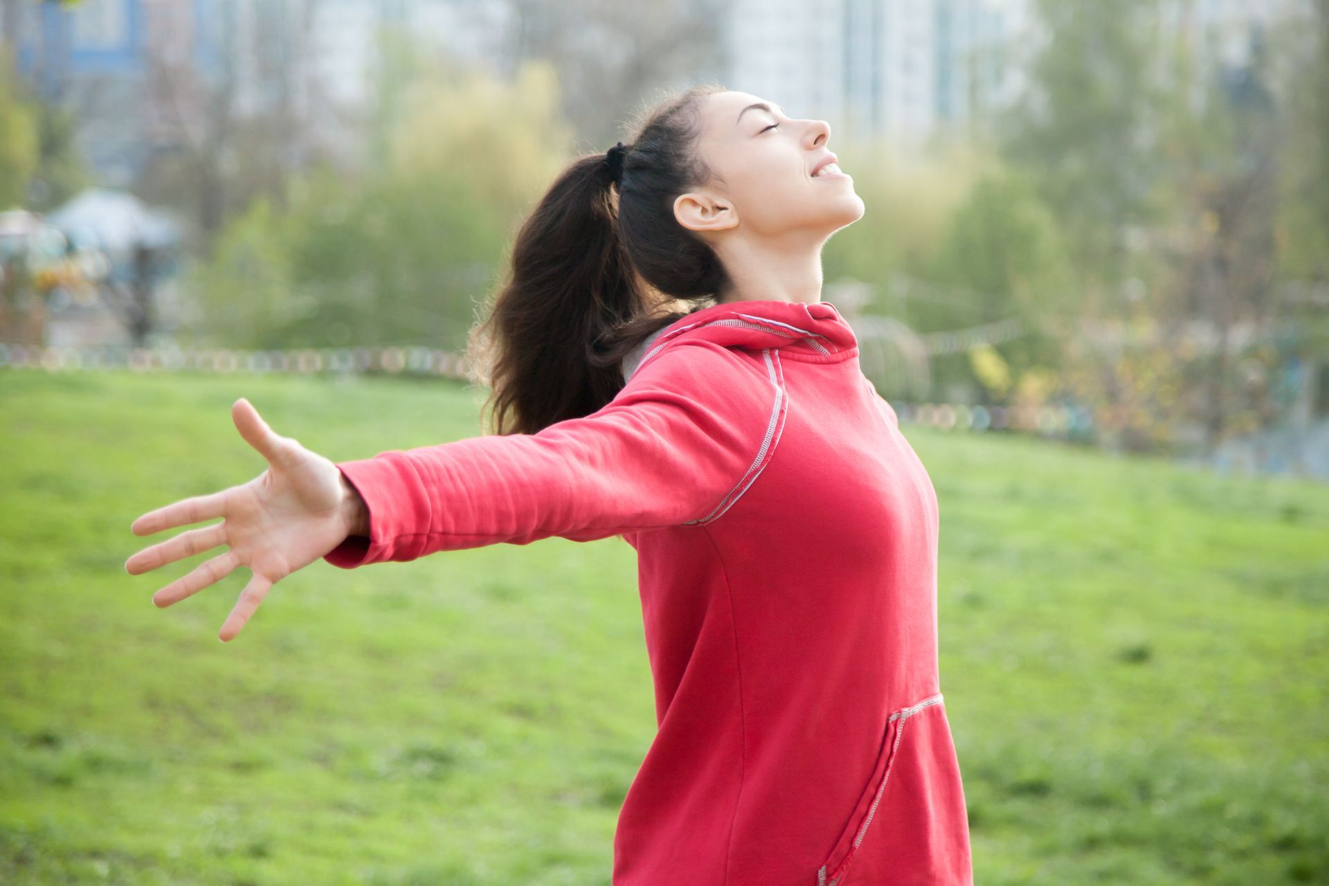 A woman in a red sweatshirt is standing in a park with her arms outstretched.